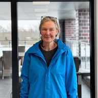Monika Huber wearing a bright blue jacket and sunglasses on her head, smiling in an indoor setting with glass doors and terrace visible behind.
