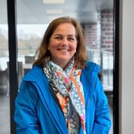 Michaela Kuhn smiling, wearing a bright blue jacket and colorful floral scarf, standing in a modern office with glass walls.