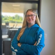 Lea Sophie Döscher mit langen blonden Haaren und Brille, trägt einen blauen Hoodie mit weißem Text auf den Ärmeln, steht in einem modernen Büro.