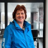 Portrait of Angelika Wagner with reddish-brown hair, smiling, wearing a bright blue jacket indoors with windows in the background.