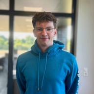 Jannik Rauchfleisch wearing glasses and a blue hoodie, standing in a modern office with glass doors and windows in the background.