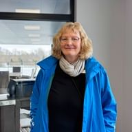 Heike Runge with blonde shoulder-length hair and glasses, wearing a blue jacket and beige scarf, standing indoors near a window.