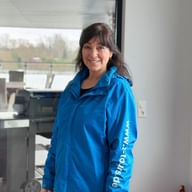 Katrin Volkmann with dark hair wearing a bright blue jacket, smiling in a modern interior with large windows showing outdoor scenery.
