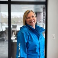 Maxi Loska wearing a bright blue jacket, smiling warmly in an indoor setting with glass doors and modern office space visible in the background.