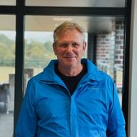 Karsten Frischknecht lächelnd in blauer Outdoorjacke und schwarzem Pullover, stehend in Innenräumen mit Fenstern und Landschaft im Hintergrund.