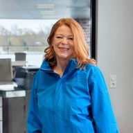Ulrike Elster with long reddish-blonde hair wearing a bright blue jacket, smiling in a modern office with glass walls and windows.