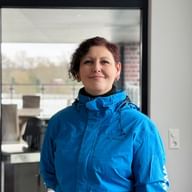 Monika Fürdos wearing a bright blue outdoor jacket, smiling in an indoor setting with large windows showing an outdoor view.