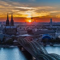 Luftaufnahme von Köln bei Sonnenuntergang mit gotischem Dom, Hohenzollernbrücke über dem Rhein und Stadtsilhouette mit dramatischen Wolken.