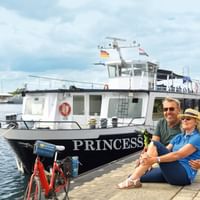 Paar mit Fahrrädern sitzt am Kai neben weißem Flusskreuzfahrtschiff MS PRINCESS. Rotes Fahrrad im Vordergrund, Marina und bewölkter Himmel im Hintergrund.