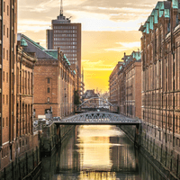 Red brick warehouse buildings line a canal in Hamburg's Speicherstadt district. A metal bridge crosses the water, reflecting golden sunset light.
