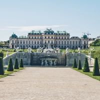 Barockes Schloss Bellevue in Wien mit formalen Gärten, weißen Statuen, gepflegten Hecken und Fontänenkaskade im Vordergrund.