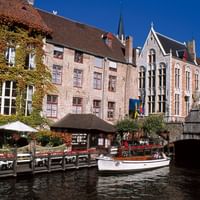 Weißes Touristenboot auf Gracht in Brügge mit mittelalterlichen Backsteingebäuden, efeubedeckten Fassaden und Steinbrücke unter blauem Himmel.