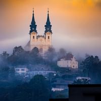 Historische Kirche mit barocken Zwillingstürmen erhebt sich über neblige Wohngebäude in Linz während der goldenen Stunde mit dramatischem Himmel.