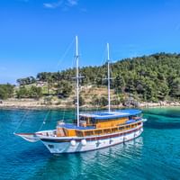 Segelyacht San Snova in türkisfarbenem Wasser Weiße Segelyacht San Snova mit zwei Masten vor Anker in kristallklarem türkisfarbenem Wasser nahe einer bewaldeten Küste unter blauem Himmel.