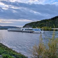 MS SE-MANON Flusskreuzfahrtschiff fährt auf dem Rhein bei Andernach mit bewaldeten Hügeln und dramatischem Wolkenhimmel bei Sonnenuntergang.
