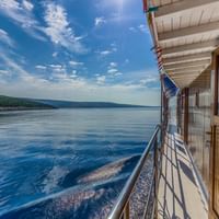 Linda Schiff Deck 3 mit Meerblick Blick von Deck 3 des Schiffes Linda mit Holzgeländer, Kabinentüren und blauem Meerwasser mit bewaldeter Küste unter bewölktem Himmel.