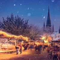 Verschneiter Weihnachtsmarkt bei Dämmerung mit beleuchtetem Karussell, geschmücktem Weihnachtsbaum, Marktständen und gotischen Kirchtürmen.