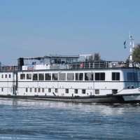 Weißes Flusskreuzfahrtschiff De Nassau fährt auf ruhigem Wasser. Das mehrstöckige Schiff hat große Fenster und ein Sonnendeck, grüne Ufer sind sichtbar.