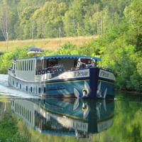 MS Fleur Flusskreuzfahrtschiff liegt an einem ruhigen Gewässer, umgeben von üppigen grünen Bäumen und Vegetation, mit perfekter Spiegelung im Wasser.