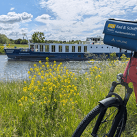 River cruise ship Casanova with blue and white hull on waterway. Red bicycle with tour company sign in foreground, yellow wildflowers along shore.