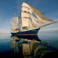 Blue-hulled sailing ship Antigua with white sails unfurled, reflected in calm turquoise waters under a clear blue sky.