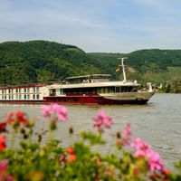 MS Serenade 1 Flusskreuzfahrtschiff fährt auf ruhigem Wasser mit bunten Blumen im Vordergrund und grünen Hügeln im Hintergrund.