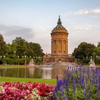 Historischer Mannheimer Wasserturm umgeben von formalen Gärten mit bunten Blumenbeeten, Spiegelteich und alten Bäumen unter bewölktem Himmel.