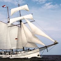 Die Flying Dutchman, ein weißes Segelschiff mit voll entfalteten weißen Segeln, segelt auf dunkelblauen Wasser unter bewölktem Himmel.