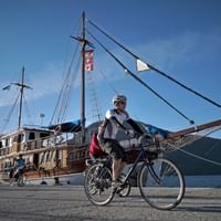Radfahrer mit Helm auf Fahrrad vor traditionellem Holzsegelschiff Panagiota im Hafen, mit Bergen im Hintergrund.