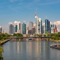 Frankfurt am Main Skyline mit modernen Wolkenkratzern, die sich im Main spiegeln. Eine Brücke überspannt das Wasser mit Booten.