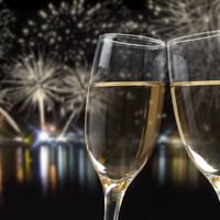 Zwei Sektgläser gefüllt mit Sekt vor einem Hintergrund aus goldenem Feuerwerk über Wasser mit reflektierten Stadtlichtern.