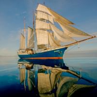 Dreimastiges Segelschiff MS Antigua mit weißen Segeln auf ruhigem blauen Wasser, das perfekte Spiegelungen unter klarem Himmel erzeugt.