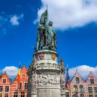 Bronzedenkmal mit zwei Figuren und Fahne am Grote Markt in Brügge, umgeben von bunten mittelalterlichen Gildenhäusern unter blauem Himmel.