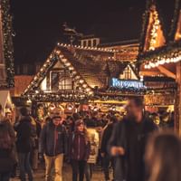 Abendszene auf einem Weihnachtsmarkt mit Holzbuden voller warmer Lichter. Besucher wandeln zwischen beleuchteten Ständen mit Essen und Geschenken.