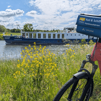 Flusskreuzfahrtschiff Casanova mit blau-weißem Rumpf auf Wasserstraße. Rotes Fahrrad mit Reiseveranstalter-Schild im Vordergrund, gelbe Wildblumen am Ufer.