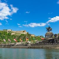 Deutsches Eck in Koblenz mit Reiterstandbild, bunten Flaggen, Rhein und Festung Ehrenbreitstein auf dem Hügel unter blauem Himmel.