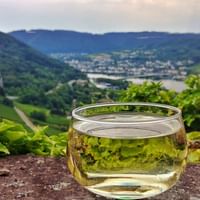 Glas Weißwein auf Steinmauer mit Panoramablick auf das Moseltal, Weinberge an Hängen und Stadt unten unter bewölktem Himmel.