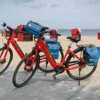 Zwei rote Fahrräder mit blauen Packtaschen auf Holzsteg am Ostseestrand mit bunten Strandkörben im Hintergrund.