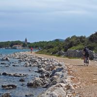 Radfahrer auf Küstenweg in Losinj, Kvarner Bucht Radfahrer auf einem Kiesweg entlang der Küste in Losinj, Kvarner Bucht. Felsige Küste mit klarem blauen Wasser links, Kirchturm in der Ferne.