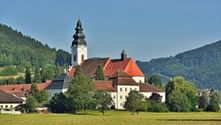 Stift Engelhartszell mit weißem Barockkirchturm und rotgedeckten Gebäuden auf grüner Wiese, umgeben von bewaldeten Hügeln.