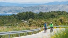 Zwei Radfahrer auf einer asphaltierten Straße bei Hvar mit Bergen, Meer und üppiger Vegetation im Hintergrund unter teilweise bewölktem Himmel.