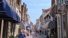 Fußgänger-Einkaufsstraße in Enkhuizen mit traditionellen holländischen Backsteingebäuden, blauen Markisen und Passanten auf Kopfsteinpflaster.