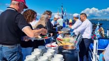 Guests being served food at an outdoor barbecue buffet on a ship deck at Stettiner Haff, with blue water and coastline visible in the background.
