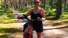 Woman with sunglasses on red bicycle on sunny forest path on Usedom. Tall pine trees line the trail with green undergrowth.