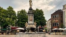 Ornate stone fountain with statue on top in Deventer town square, surrounded by historic buildings, cafes with outdoor seating and green trees.