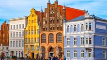 Row of colorful historic buildings in Stralsund with Gothic brick architecture. People gather in the square in front of white, yellow, red-brick, and blue facades.