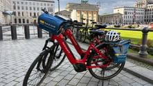 Rote und gelbe Leihräder auf Kopfsteinpflaster mit dem Brandenburger Tor im Hintergrund unter bewölktem Himmel in Berlin.