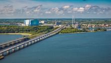 Aerial view of Rügendamm bridge crossing blue water toward Stralsund. Green forests line the shores, with industrial buildings and city visible beyond.