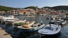 Hafen von Pučišća auf Brač mit vertäuten Booten im Vordergrund, Steingebäuden mit roten Dächern und Kirchturm. Grüne Hügel umgeben die Stadt.