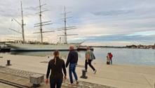 People walking along the harbor promenade in Stralsund with a white three-masted sailing ship moored at the quay under a cloudy sky.